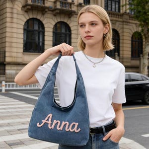 May include: A denim tote bag with the name "Anna" embroidered in pink script. The bag has long straps and a button closure. The background features a city street and a building.