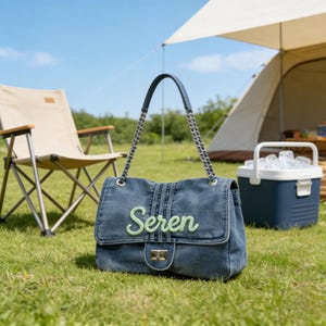 May include: A denim handbag with a silver chain strap and a flap closure. The word "Seren" is embroidered in green on the front. The bag is sitting on green grass, with a camping chair and tent in the background.
