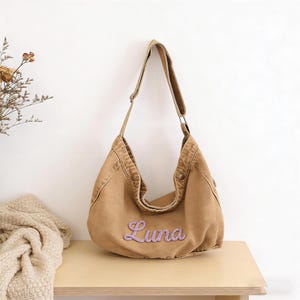 May include: A tan-colored shoulder bag with the name "Luna" embroidered in lavender script. The bag has a wide strap and a curved design. It is made of a sturdy fabric and is displayed on a light-colored wooden surface.