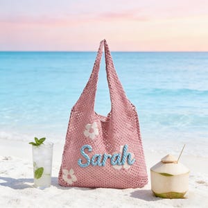 May include: A pink crocheted tote bag with the name "Sarah" in blue script, decorated with white flower appliqués. The bag is on a sandy beach with a cocktail and a coconut. The ocean and sky are in the background.