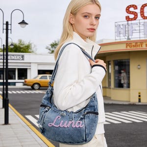 May include: A denim shoulder bag with the name "Luna" embroidered in pink script. The bag has a zipper closure and a drawstring detail. The strap is a light blue denim with a white stripe. The person is wearing a white jacket.