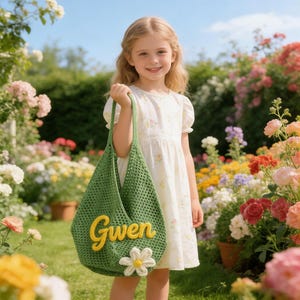 May include: A green crochet tote bag with the name "Gwen" in yellow script and a white daisy applique. The bag is held by a young girl wearing a white floral dress, standing in a garden filled with colorful flowers.