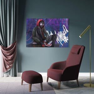 May include: A canvas print featuring a character in a red hooded jacket and black suit, with a mask, sitting against a graffiti-covered background. The artwork is displayed above a burgundy armchair and ottoman.
