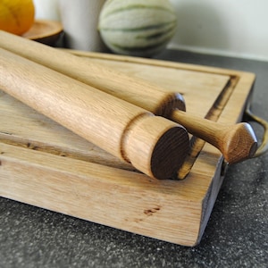 May include: A light brown wooden rolling pin with a cylindrical body and a tapered handle resting diagonally across a rectangular wooden cutting board. The rolling pin features a smooth, natural wood grain finish and a distinct circular end cap.