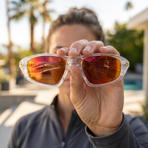 K&ouml;nnte beinhalten: Eine Person h&auml;lt eine Sonnenbrille mit transparentem Rahmen und rot-orangen Gl&auml;sern vor ihr Gesicht. Die Gl&auml;ser spiegeln die Umgebung wider. Die Sonnenbrille hat eine rechteckige Form und kleine goldene Akzente an den B&uuml;geln.