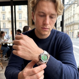 Könnte beinhalten: Eine silberfarbene Armbanduhr mit grünem Zifferblatt. Die Uhr hat ein strukturiertes Metallarmband und ein rundes Zifferblatt mit einem einzigartigen Design. Die Uhr wird am Handgelenk getragen.