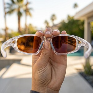 K&ouml;nnte beinhalten: Eine Sonnenbrille mit transparentem Rahmen und orange get&ouml;nten Gl&auml;sern wird vor einem verschwommenen Hintergrund im Freien gehalten. Die Sonnenbrille hat eine rechteckige Form und kleine goldfarbene Akzente in der N&auml;he der Scharniere.