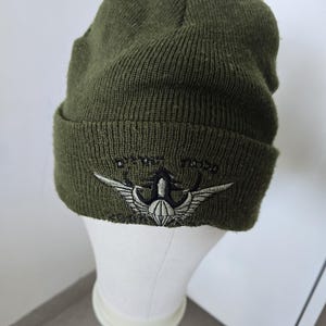 Peut inclure: Un bonnet en tricot vert olive avec un revers replié. L'avant du revers présente un motif brodé d'une ancre ailée argentée avec des détails noirs. Le tissu tricoté a un aspect texturé et le bonnet est présenté sur un support blanc.