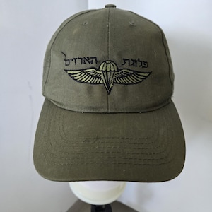 Peut inclure: Une casquette de baseball vert olive avec un emblème brodé représentant un parachute ailé au centre. Au-dessus de l'emblème, du texte hébreu noir est cousu sur le panneau avant. La casquette possède une visière courbée et une calotte structurée.