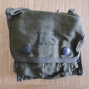 Puede incluir: Una bolsa utilitaria de lona color verde oliva con una solapa frontal asegurada por dos botones de presión de metal oscuro. Las letras U.S. están impresas en tinta negra en el centro de la solapa. La bolsa tiene una forma rectangular estructurada.