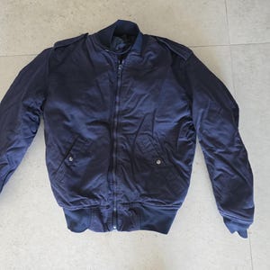 Puede incluir: Una chaqueta tipo bomber de color azul marino oscuro con cremallera frontal completa, cuello acanalado, puños acanalados y dobladillo acanalado. La chaqueta incluye dos bolsillos delanteros con cierre de botón a presión y charreteras en los hombros.