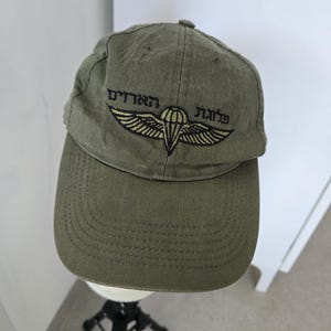 Puede incluir: Una gorra de béisbol de color verde oliva con un emblema bordado de un paracaídas con alas en hilo negro y dorado. Sobre el emblema, hay texto en hebreo bordado en negro. La gorra tiene una visera curva y una corona estructurada con botón.