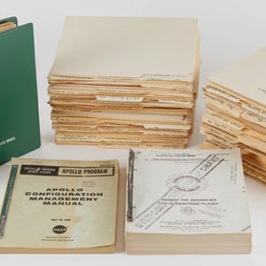 Può includere: Una collezione di documenti e manuali d'epoca relativi al programma spaziale Apollo. Include un raccoglitore verde con la scritta "Apollo Hardware Indenture", pile di cartelle e manuali con testo come "Apollo Configuration Management Manual".