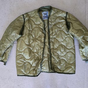 May include: An olive green quilted liner jacket featuring a wave-patterned stitch design, long sleeves, and a dark green bound edge. A white rectangular label with black text is sewn into the interior collar area of this lightweight garment.
