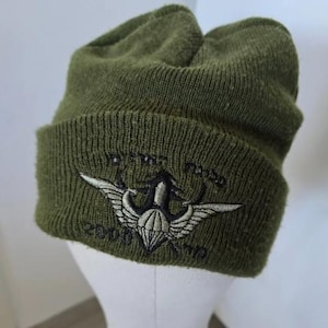 May include: Olive green knit beanie with embroidered emblem. The emblem features a parachute design with wings and text. The hat is made of a ribbed knit material and has a folded brim.