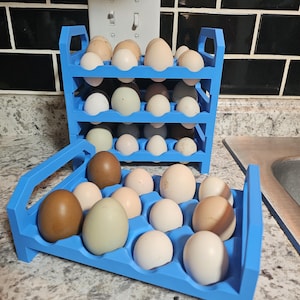 May include: A blue, three-tiered egg holder filled with various colored eggs. The eggs range in color from white to brown and green. The holder is made of a light blue material and is sitting on a countertop.