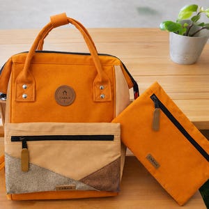 Puede incluir: Una mochila naranja con una bolsa a juego y una bolsa pequeña con cremallera. La mochila tiene un asa superior y correas para los hombros. Las bolsas tienen la marca "CABAIA". Las bolsas están hechas de un material texturizado.