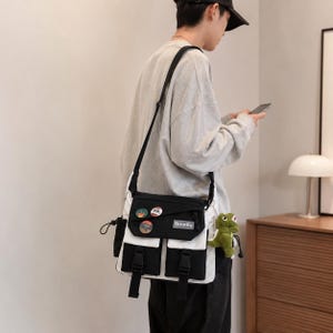 以下が含まれることがあります： 黒と白のメッセンジャーバッグ。前面に2つのポケットとフラップクロージャーが付いています。バッグには黒いショルダーストラップと前面に装飾的なピンが付いています。小さな緑色のぬいぐるみがおまけで付いています。バッグには「Binnilu」のブランド名があります。