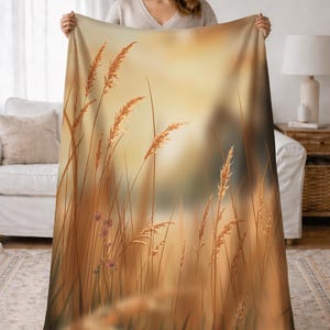 Könnte beinhalten: Eine beige Decke mit einer detaillierten Illustration von hohen, goldenen Weizenähren und Gräsern vor einem weichen, verschwommenen Hintergrund. Das Design der Decke erinnert an eine warme, natürliche Ästhetik, ideal für die Inneneinrichtung.