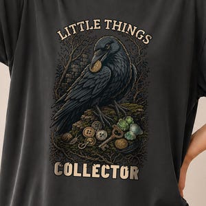 Könnte beinhalten: Ein anthrazitfarbenes T-Shirt mit einer Grafik eines schwarzen Raben, der auf einem Ast sitzt und von kleinen Gegenständen wie Knöpfen und Schlüsseln umgeben ist. Der Text über dem Raben lautet LITTLE THINGS und darunter COLLECTOR in beiger Schrift.
