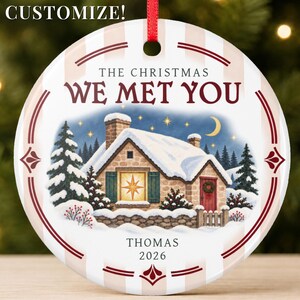 Könnte beinhalten: Ein runder, weißer Keramik-Weihnachtsbaumschmuck mit rotem Band, bedruckt mit "THE CHRISTMAS WE MET YOU", "THOMAS" und "2026". Die Illustration zeigt ein verschneites Steinhäuschen bei Nacht mit beleuchtetem Fenster, Tannenbäumen und einer Mondsichel.
