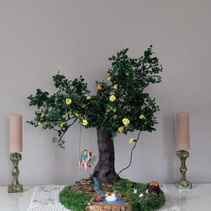 Puede incluir: Una escultura de árbol en miniatura con una pareja en un columpio, rodeada de un estanque y flores. El árbol tiene hojas verdes y rosas amarillas. Dos velas rosas se colocan a cada lado de la escultura.