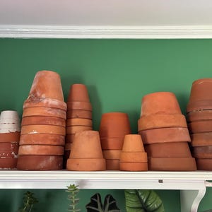 May include: A collection of terracotta flower pots of various sizes stacked on a white shelf against a green wall. The pots range in color from light to dark orange-brown, with some showing signs of age and wear.