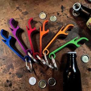 Puede incluir: Una colección de abridores de botellas de colores azul, morado, rojo, naranja y verde, cada uno con un llavero. Los abridores están colocados sobre una superficie de madera, con varias tapas de botellas y una botella de vidrio oscuro.