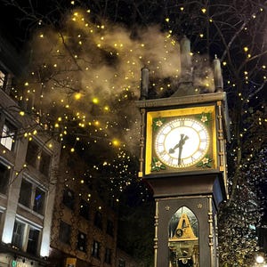 May include: A black clock tower with a white clock face and gold accents. The clock is surrounded by a wreath of flowers. The tower is lit up with string lights and steam is coming from the top.