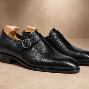 Könnte beinhalten: Ein Paar schwarze Lederschuhe mit Monkstrap-Verschluss und strukturierter Oberfläche. Die Schuhe haben einen silbernen Schnallenverschluss und werden mit hölzernen Schuhspannern präsentiert. Auf neutralem Hintergrund.