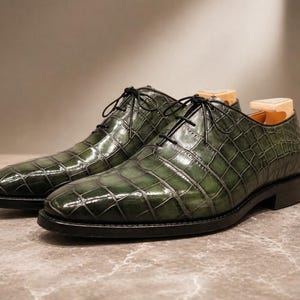 Può includere: Un paio di scarpe eleganti in pelle verde con motivo a pelle di coccodrillo. Le scarpe hanno lacci e suole neri. Le scarpe sono esposte su una superficie di marmo.