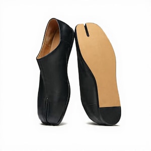 Pode incluir: Sapatos pretos de couro com biqueira dividida. Um sapato está em pé, mostrando o perfil lateral e a parte superior aberta, enquanto o outro está invertido, revelando a sola castanha e o design de biqueira dividida. Design minimalista.