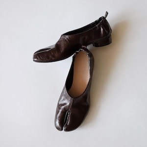 May include: Two brown leather split-toe shoes. The shoes have a low heel and a unique design with a split between the big toe and the other toes. The leather appears to be smooth and has a slight sheen.