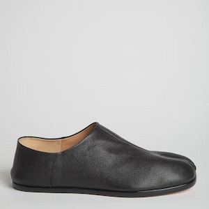 May include: A black leather slip-on shoe featuring a distinct split-toe design, a low-profile silhouette, a smooth rounded heel counter, and a central seam running from the collar to the toe box, finished with a flat, dark, low-profile sole.