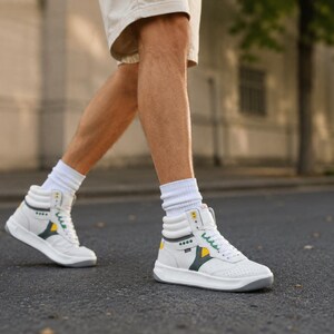 Puede incluir: Zapatillas altas blancas con detalles amarillos y verdes. Las zapatillas tienen una suela gris y se usan con calcetines blancos. Los zapatos est&aacute;n sobre una superficie de asfalto gris oscuro.