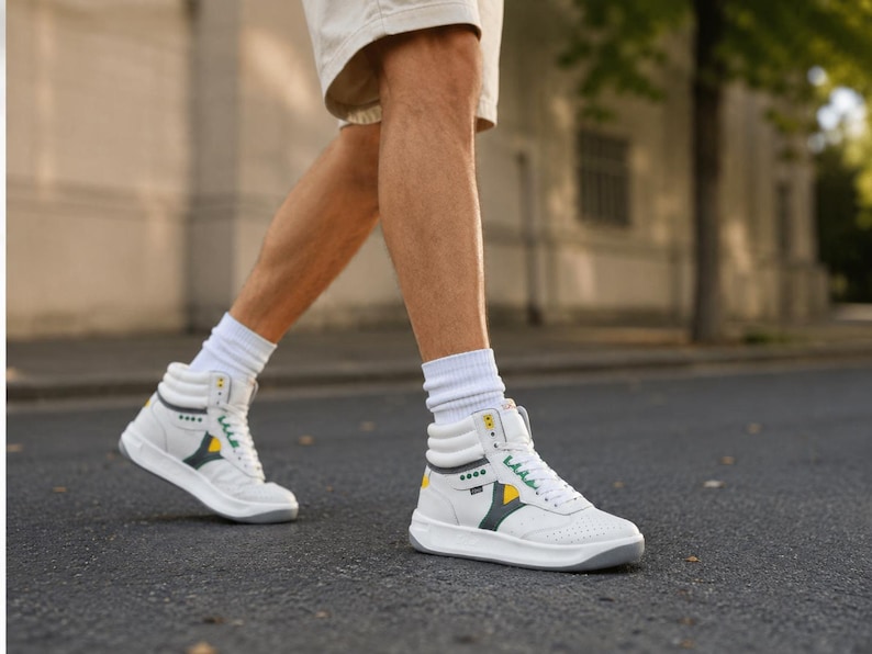 Puede incluir: Zapatillas altas blancas con detalles amarillos y verdes. Las zapatillas tienen una suela gris y se usan con calcetines blancos. Los zapatos est&aacute;n sobre una superficie de asfalto gris oscuro.