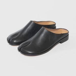 May include: A pair of black leather slip-on shoes with a split-toe design. The shoes have a low heel and a smooth, matte finish. The design is reminiscent of traditional Japanese tabi socks.