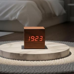 May include: A wooden cube digital clock displaying 19:23 in red numerals. The clock sits on a white marble-like stand, placed on a white rug. The background shows a bed with white linens and a side table.