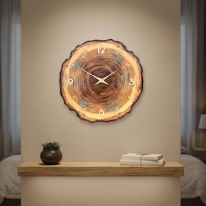 May include: A circular wooden wall clock with a tree ring pattern. The clock has white numerals at 12, 3, 6, and 9 o'clock, and gold-coloured hands. It is mounted on a beige wall, suitable for a living room or bedroom.