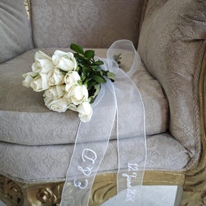 Puede incluir: Un ramo de rosas blancas con hojas verdes y una cinta blanca transparente. La cinta está bordada con las iniciales "E & D" y la fecha "12 de junio de 2016". El ramo descansa sobre una silla tapizada beige estampada.