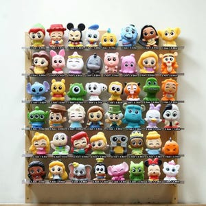 Puede incluir: Una colección de coloridas figuras de personajes, cada una de aproximadamente 2,5 cm de altura, expuestas en estantes transparentes. Las figuras representan varios personajes de dibujos animados, incluyendo Mickey Mouse y Stitch. La exhibición está montada sobre un fondo de madera.