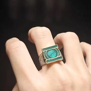 May include: A silver-toned metal ring featuring a rectangular face with a central circular emblem. The emblem and rectangular border are filled with a textured, shimmering green inlay, framed by thin gold-colored metal edges.