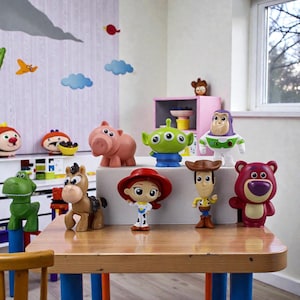May include: A collection of Toy Story character figurines, including Woody, Buzz Lightyear, and Jessie, arranged on a wooden table. The figures are brightly colored and detailed, with a pink pig, a green alien, and a brown horse also present. The background features decorative wall art.