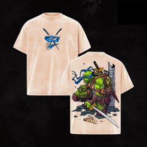 May include: Tan-colored t-shirt featuring a blue ninja turtle design on the front and back. The front displays crossed swords and a blue mask. The back showcases a ninja turtle with a sword, set against a cityscape backdrop.