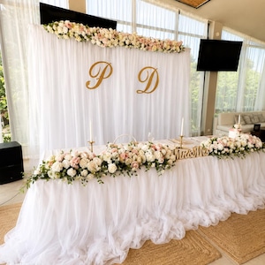 Può includere: Un tavolo e uno sfondo coperti di tulle bianco, decorati con composizioni floreali di rose bianche e rosa. Lo sfondo presenta grandi lettere dorate in corsivo P e D. Il tavolo include due candele alte e sottili e un cartello in legno.