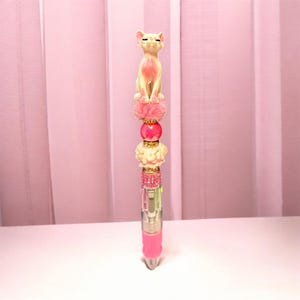 Puede incluir: Un bolígrafo decorativo con una figura de gato de color rosa claro esculpida en la parte superior, seguido de un pompón rosa esponjoso, una cuenta rosa intenso lisa, una cuenta texturizada de color crema y una banda de pequeños diamantes de imitación rosas sobre un cuerpo transparente y rosa.