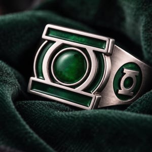 Pode incluir: Um anel de metal prateado com uma face retangular proeminente, uma pedra verde circular central e detalhes em esmalte verde. A lateral do anel exibe um emblema circular com fundo verde e um símbolo de lanterna prateado.