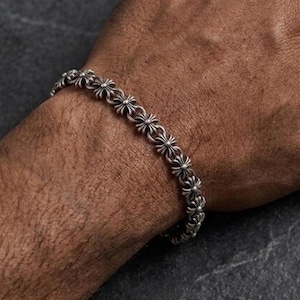 May include: A silver bracelet featuring a repeating floral pattern. The bracelet is composed of small, interconnected flower-shaped links. The bracelet is worn on a wrist, and the metal has a slightly aged appearance.