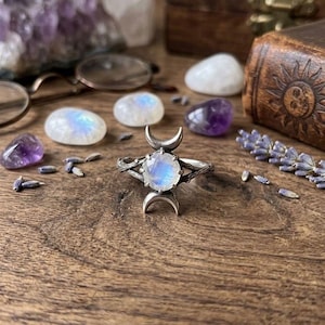 May include: A silver ring featuring a round, iridescent moonstone cabochon at the center, flanked by two crescent moon shapes. The band has a textured, branch-like appearance, and the metal has a dark, oxidized finish to highlight the details.