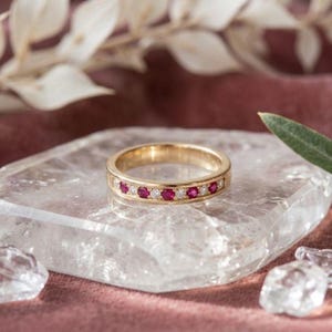 May include: A gold band ring with alternating ruby and diamond gemstones. The ring is set on a clear, crystalline surface, with a soft pink background. The ring's design features a row of vibrant red rubies and sparkling diamonds.